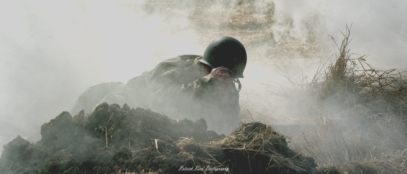 "Image of a soldier standing resolutely in a smoky field, surrounded by a haze of battle. The soldier, dressed in a WWII-era uniform, grips his rifle tightly, scanning the environment with a determined expression. The smoke curls around him, creating a dramatic atmosphere that highlights the chaos of war. The background features silhouettes of trees and hints of distant action, emphasizing the soldier's isolation and bravery in a challenging situation."